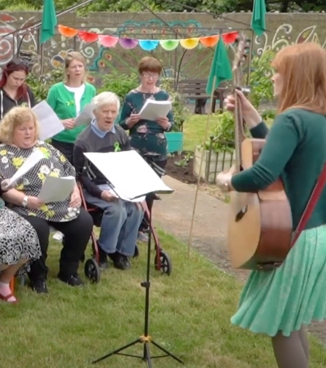Men and women in a garden singing with a girl dressed in green playing the guitar
