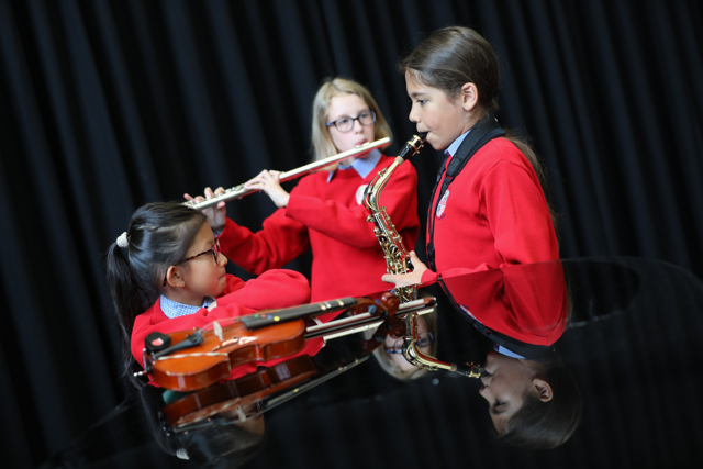 Three young girls in red jumpers play instruments at a grand piano