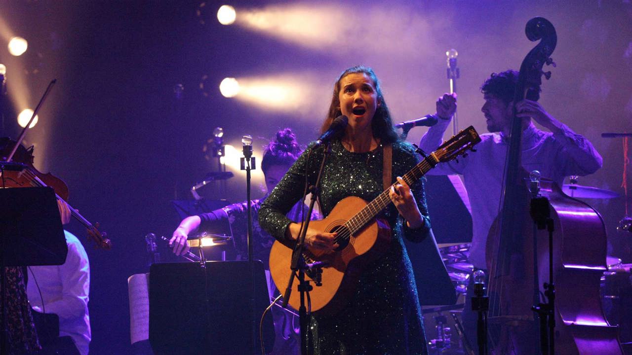 Singer Lisa Hannigan singing with a guitar in a purple light with additional musicians in the background
