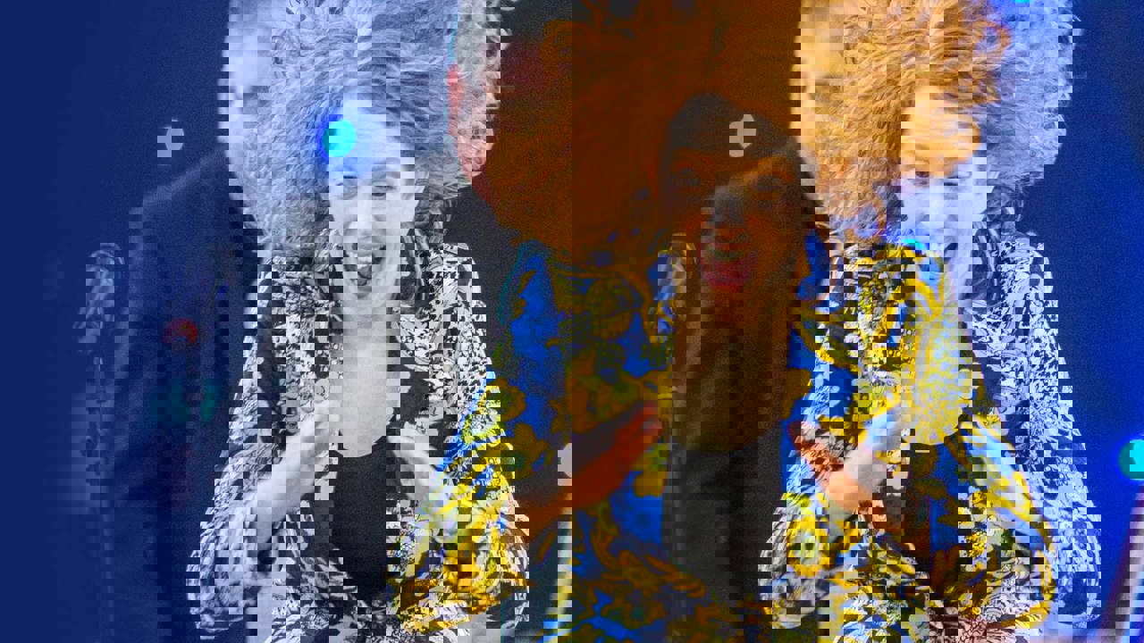 Jessie Grimes - woman in a blue and yellow paisley jacket with red hair in motion against a blue backdrop