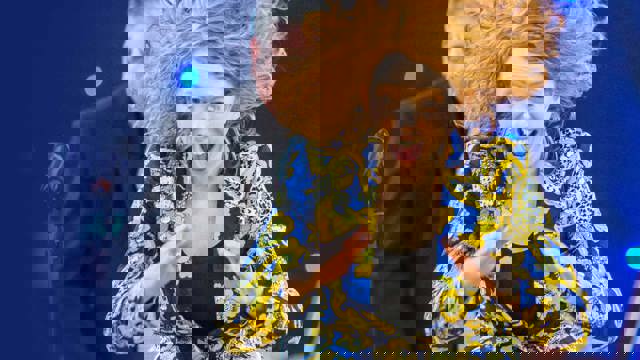 Jessie Grimes - woman in a blue and yellow paisley jacket with red hair in motion against a blue backdrop