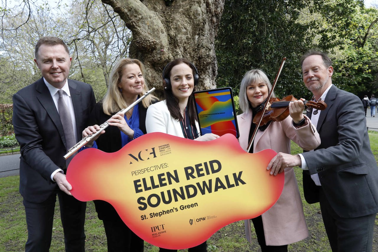 Five people in a park; two men holding a sign saying Ellen Reid Soundwalk with two women playing a flute and violin while another woman holds a mobile device