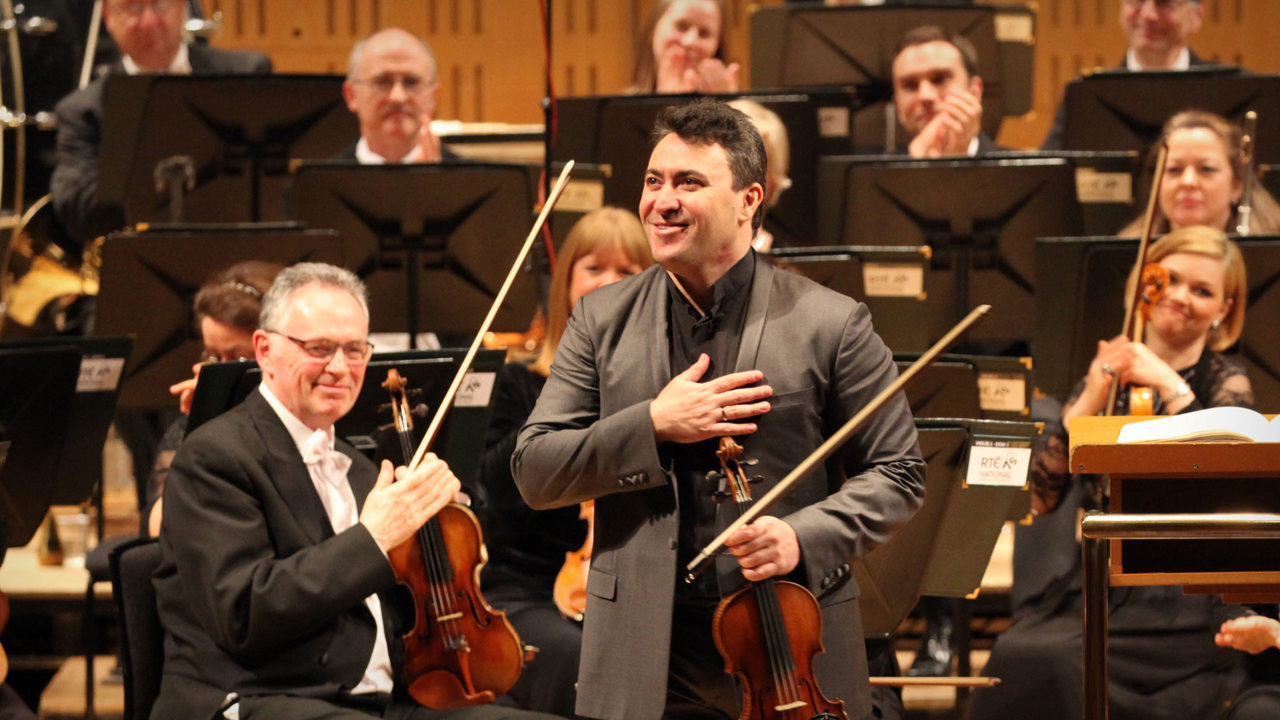 Maxim Vengerov with his violin and hand on his chest taking a bow with members of the National Symphony Orchestra in the background