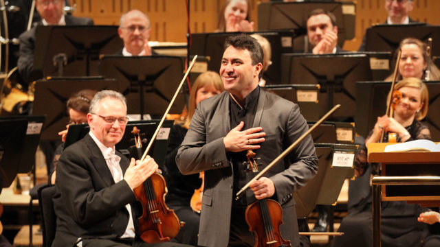 Maxim Vengerov with his violin and hand on his chest taking a bow with members of the National Symphony Orchestra in the background