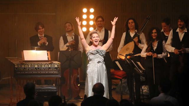 Mezzo-soprano Joyce DiDonato in a silver dress with her arms in the air with Il Pomo D'oro in the background at curtain call