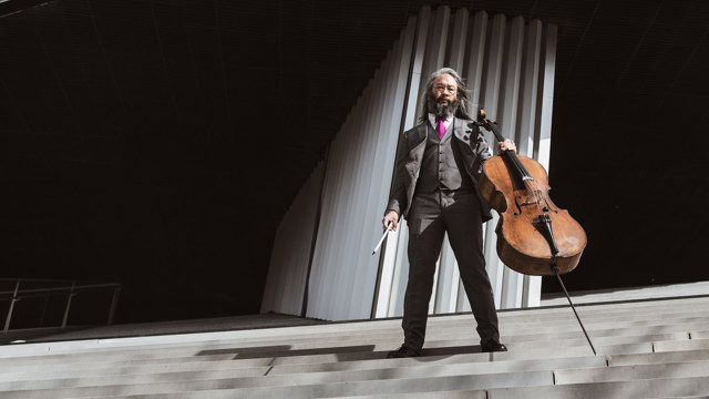 Éric Maria Couturier - man on grey steps in a grey suit with a cello and bow