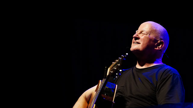 Christy Moore - man in black with a guitar against a black background