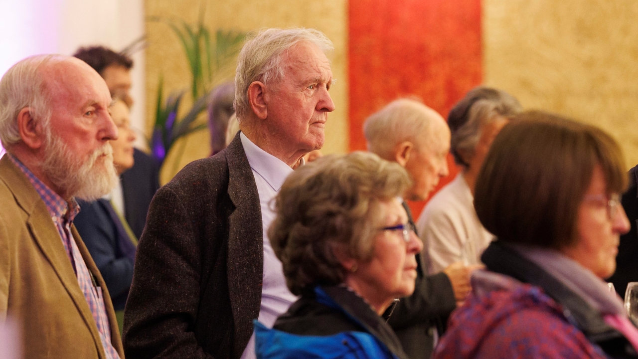 Audience members, male & female, looking at something in the distance - some in focus, some are out of focus