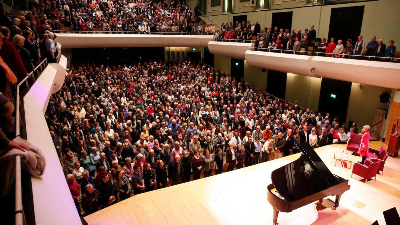 Wide shot of a full house from Choir Balcony of NCH stage during a standing ovation with a grand piano and chairs on stage